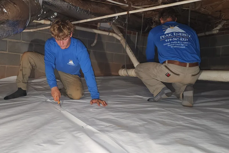 Two Peak Energy crew members trimming and seaming the white vapor barrier liner on a crawl space floor in NC