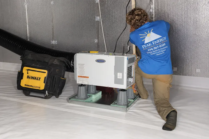 Peak Energy technician setting an Aprilaire dehumidifier onto its risers in a finished encapsulated crawl space with white vapor barrier walls and floor in NC