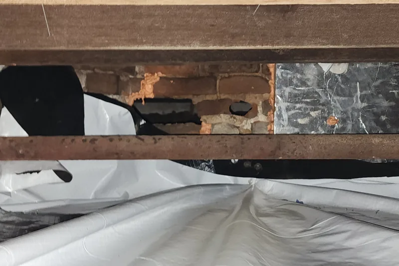 Crawl space liner left torn and unsealed at the rim joist with an open gap to bare brick in an NC home