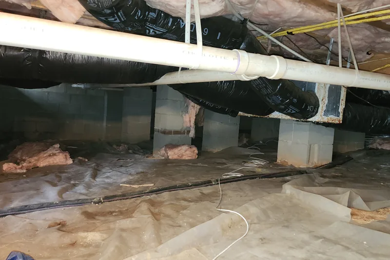Torn, shifted ground vapor barrier in a vented crawl space with bare soil exposed between foundation piers