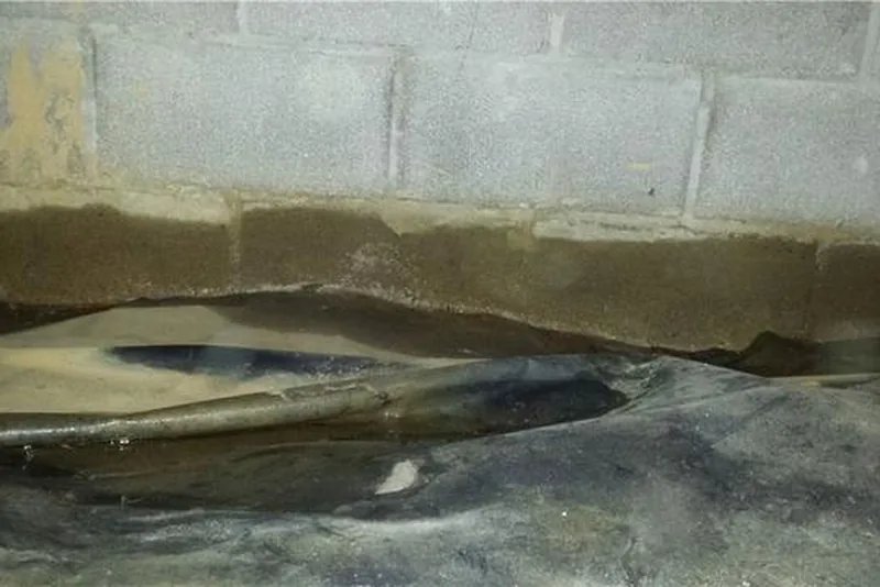 Water staining along the base of a concrete block foundation wall above a dark, damp vapor barrier in a crawl space