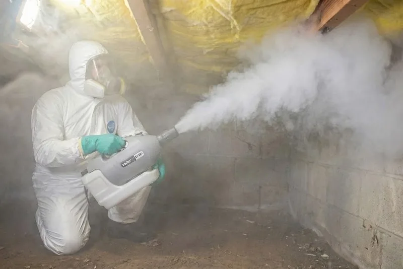 Technician in protective suit and respirator fogging an antimicrobial treatment inside a crawl space with a block foundation wall and insulation overhead