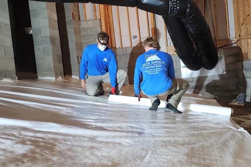 Peak Energy crew rolling out a white polyethylene vapor barrier across a crawl space floor in a Wake County NC home