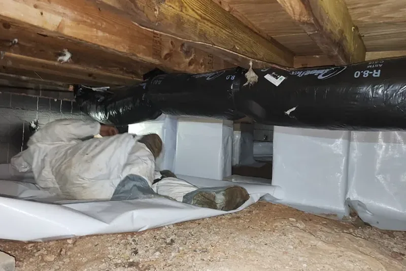 Peak Energy crew member in protective coveralls installing a heavy-duty white vapor barrier across a crawl space floor in Wake County NC, with wrapped piers and insulated ductwork already in place