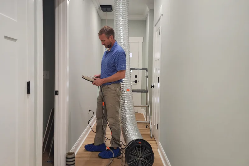 Peak Energy technician reading a digital pressure gauge while a duct blaster fan runs in a finished home hallway in Wake County NC, checking the test result