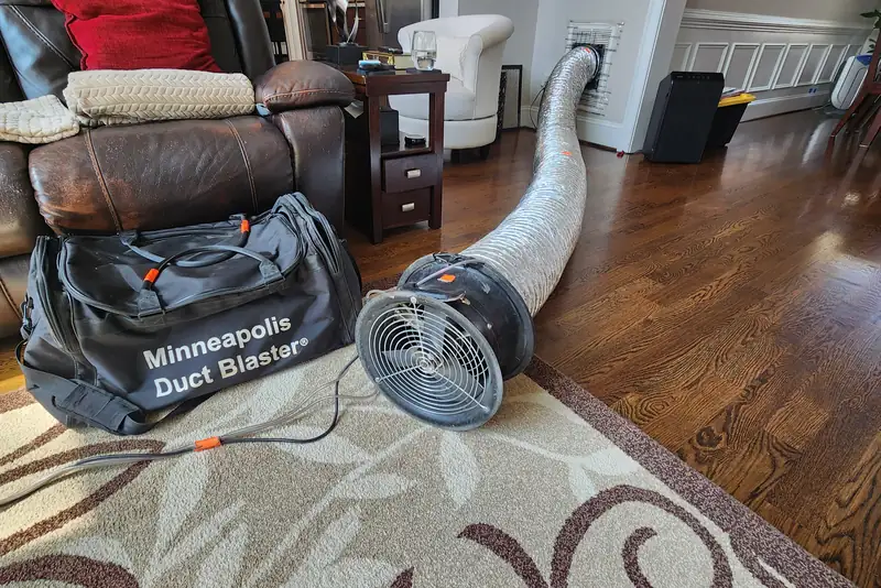 Minneapolis Duct Blaster fan and equipment bag connected by flex duct to a wall vent in a furnished Wake County NC living room, set up for a duct leakage test as part of a home energy audit