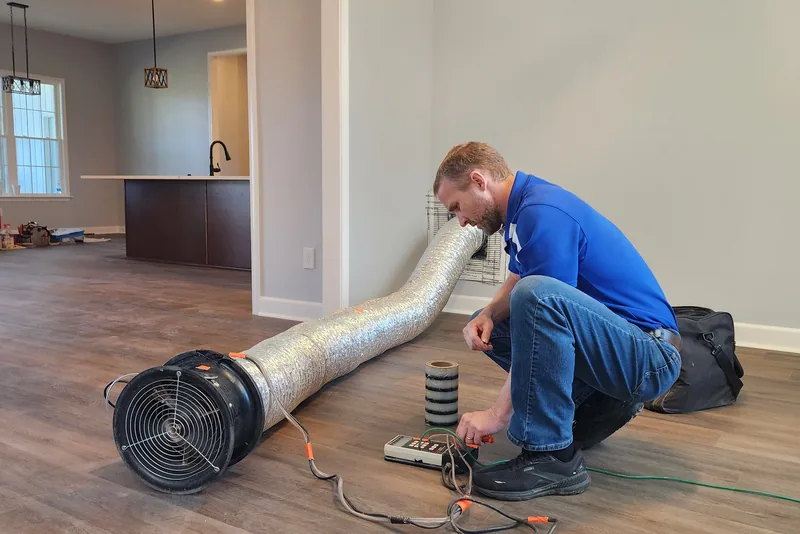 Peak Energy technician setting up a duct blaster fan connected to a return air grille in a new construction home in Wake County NC, with digital pressure gauge on the floor