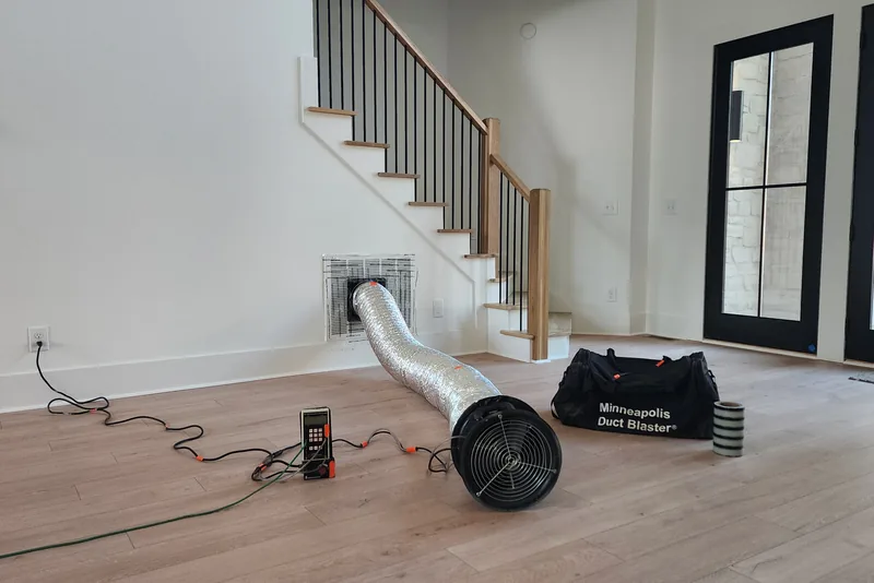 Minneapolis Duct Blaster fan connected to a floor return register with flexible duct, manometer on the floor beside it, during duct leakage testing in a Triangle NC home