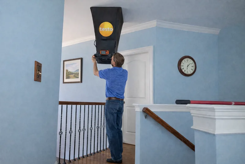 Peak Energy technician measuring supply airflow at a ceiling register with a Testo flow hood during a home energy audit in a Triangle NC home