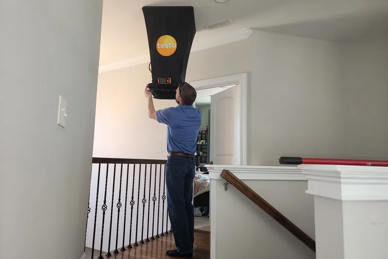 Peak Energy technician holding a Testo flow hood up to a bedroom supply register to measure delivered airflow during a home energy audit in a Triangle NC home