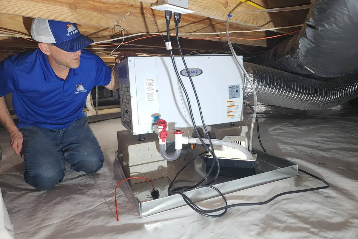 Peak Energy technician installing an AprilAire dehumidifier in an encapsulated crawl space in Holly Springs NC — white vapor barrier on floor, conditioned ductwork in background