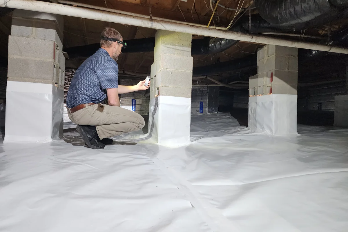 Peak Energy technician with headlamp inspecting an encapsulated crawl space in the Triangle NC — sealed piers, white vapor barrier, conditioned ductwork